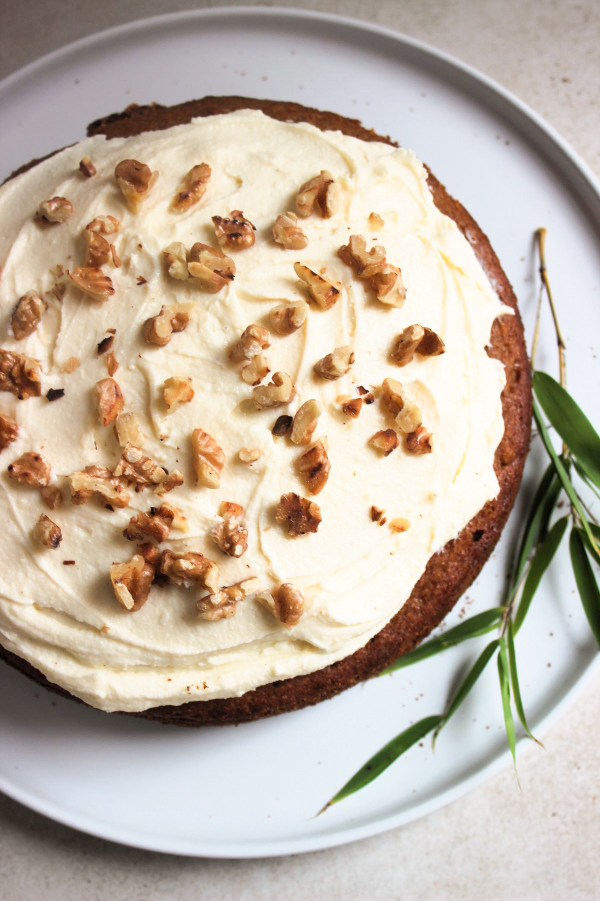 Sweet potato cake with cream cheese frosting and chopped nuts on a white plate seen from above. A twig with leaves on the side of the cake.