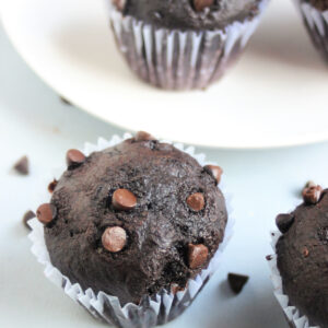 Double chocolate muffin in a liner on a light blue surface. More muffins behind.