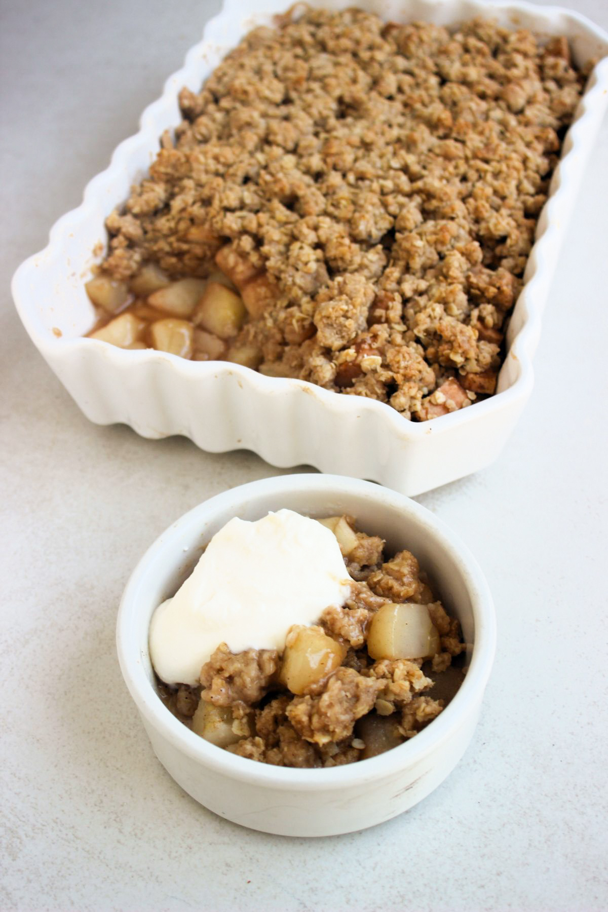 A small pot with pear crisp with whipped cream. Behind, a rectangular baking dish with pear crisp.