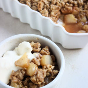 Tiny bowl with pear crisp and whipped cream. Baking dish behind with pear crisp.