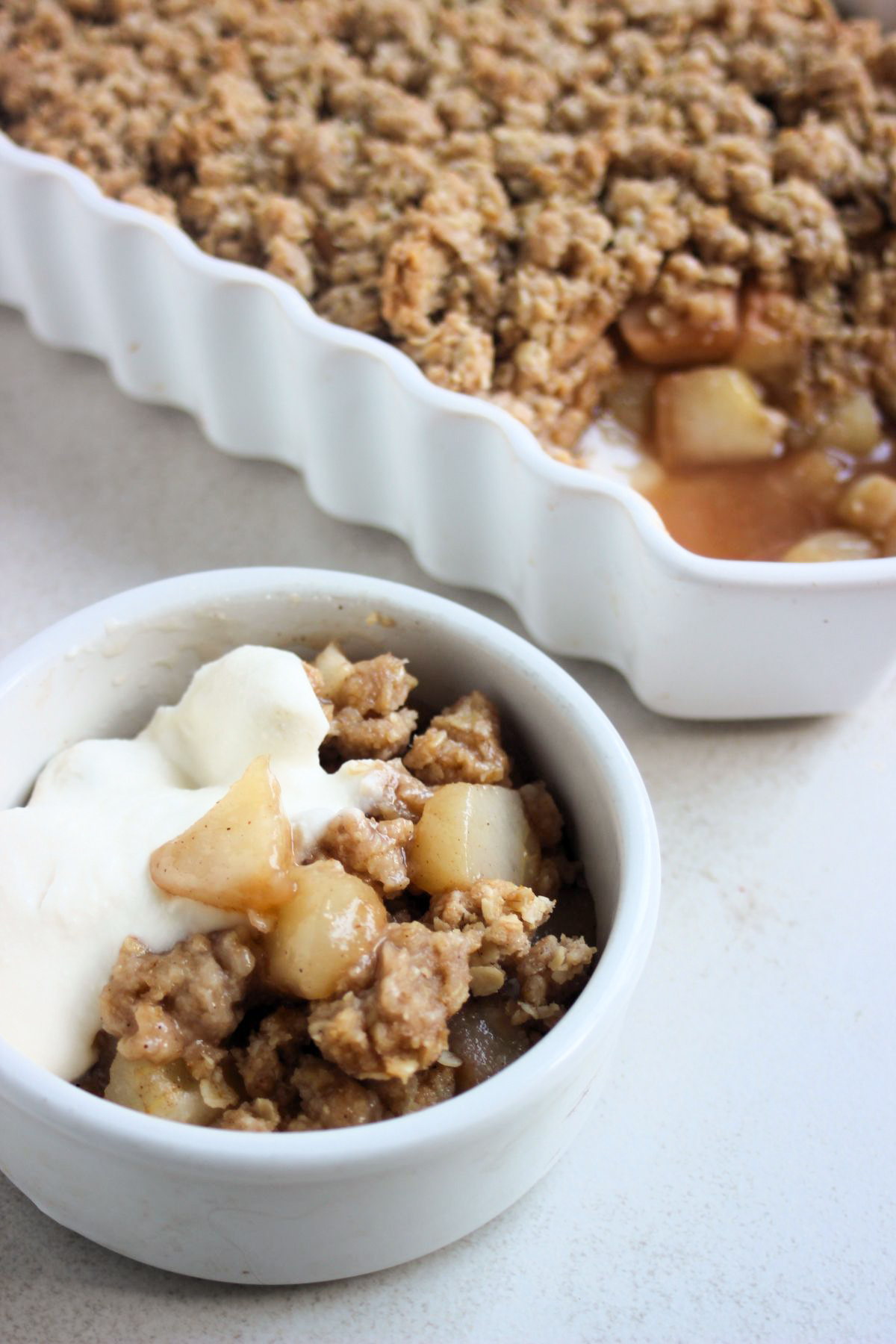 Tiny bowl with pear crisp and whipped cream. Baking dish behind with pear crisp.