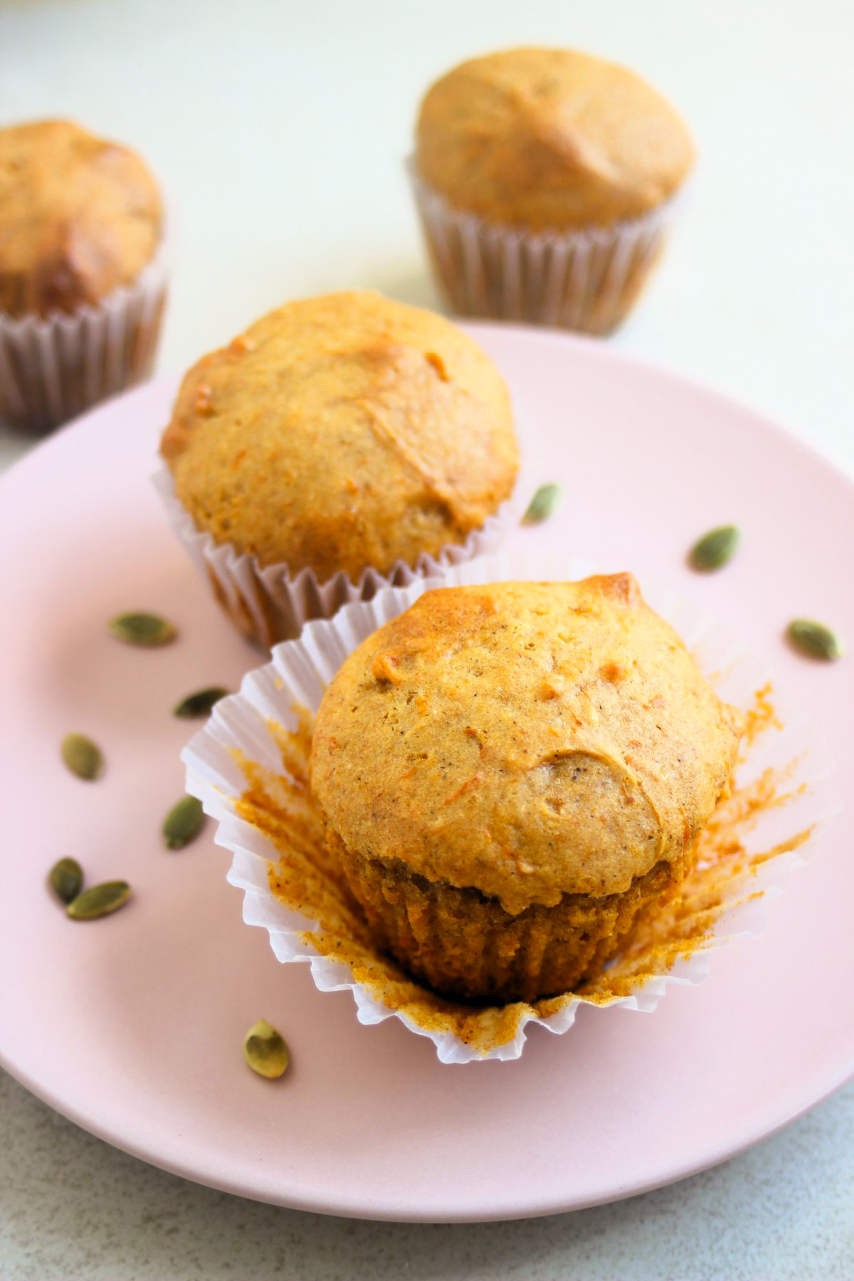 Pumpkin muffin with the liner open and another muffin behind on a pink plate. Pumpkin seeds are scattered on the plate.