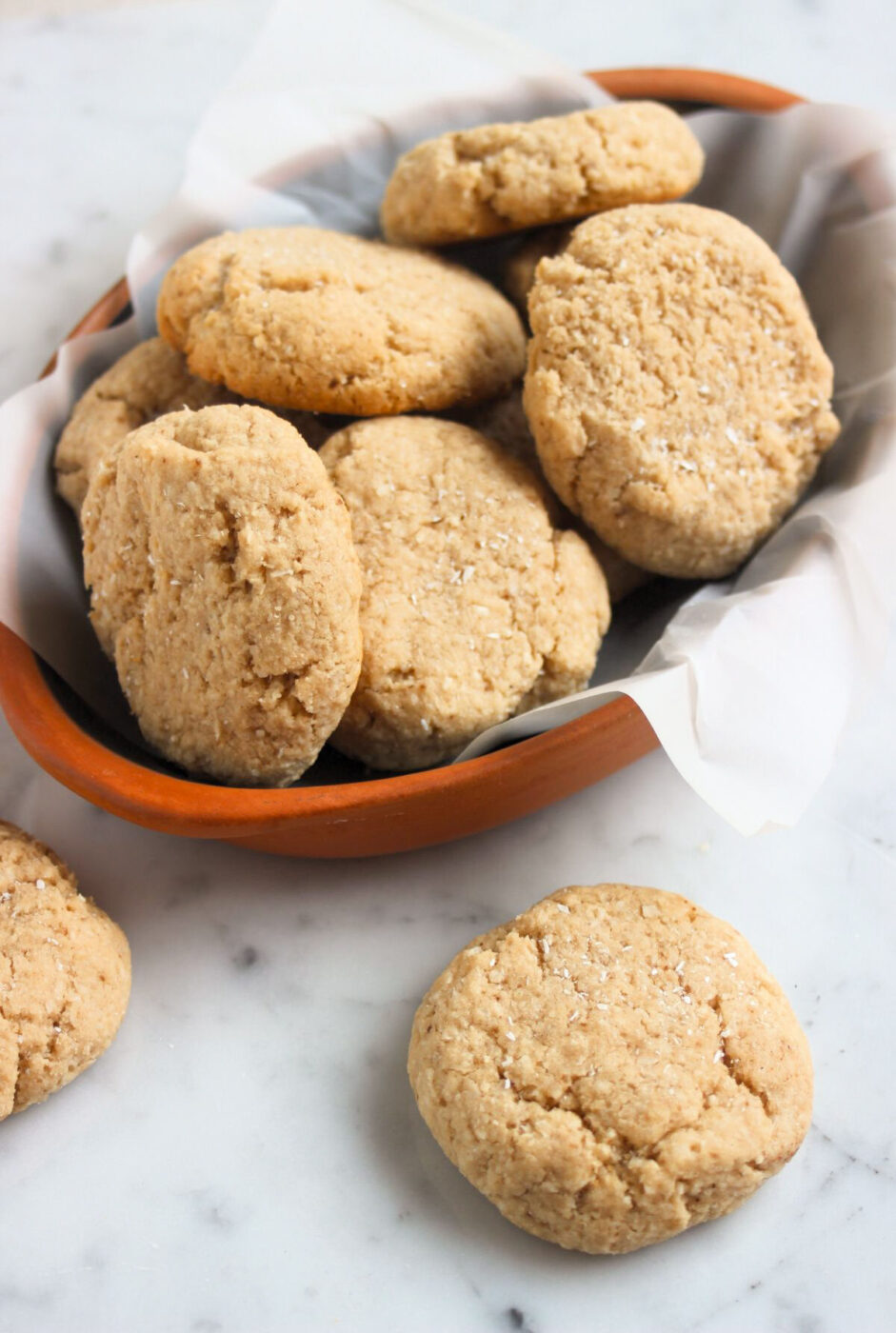 Coconut cookies on a white surface and more cookies behind in a deep dish.