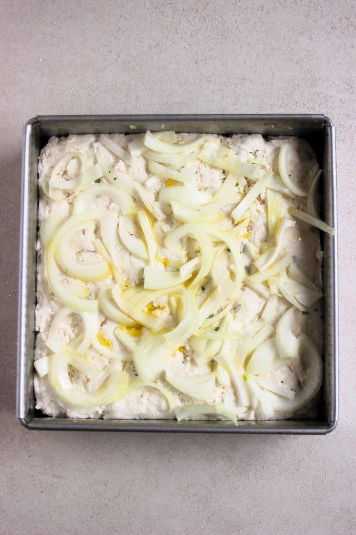 Square baking pan with focaccia dough with onion slices and oil.