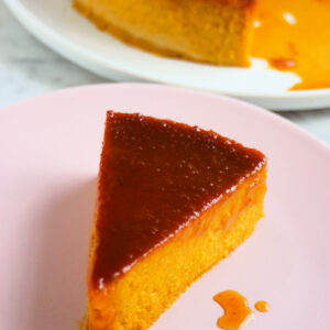 A portion of pumpkin flan with caramel on a pink plate. Whole flan behind.