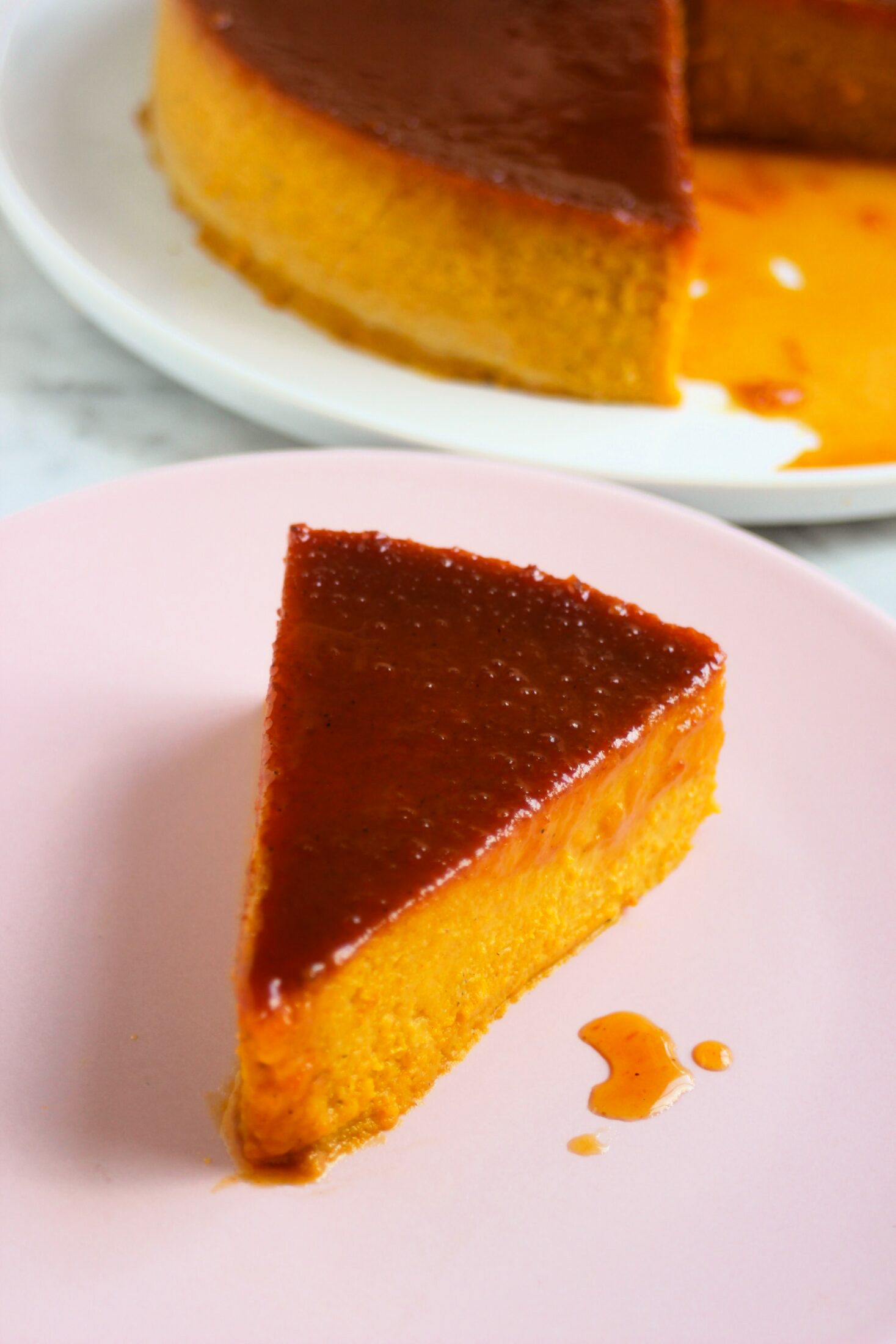 A portion of pumpkin flan with caramel on a pink plate. Whole flan behind.